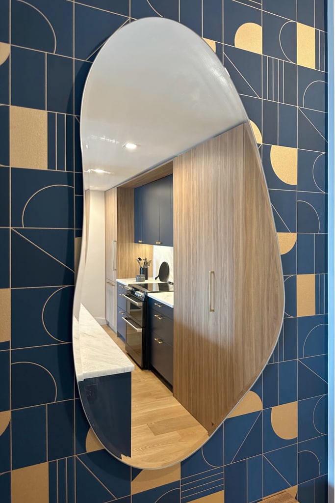 View of the wall covered in a custom wallpaper with a tiled pattern in deep blue and bronze. The curved mirror reflects the galley kitchen using the same color palette.